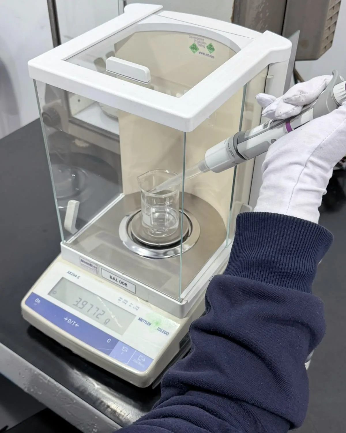 Mão com luva pipeta água num béquer dentro da balança analítica, para calibrar a micropipeta