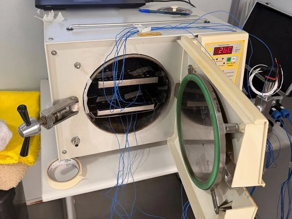 Autoclave de bancada com a porta aberta e os termopares azuis do ensaio passando pelas bandejas, com o indicador marcando 26 °C.