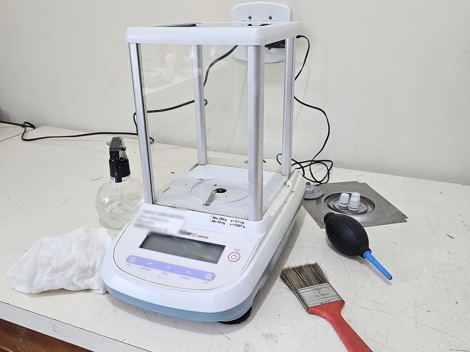 Balança analítica limpa na bancada, com pincel, soprador de ar e pano usados na manutenção preventiva.